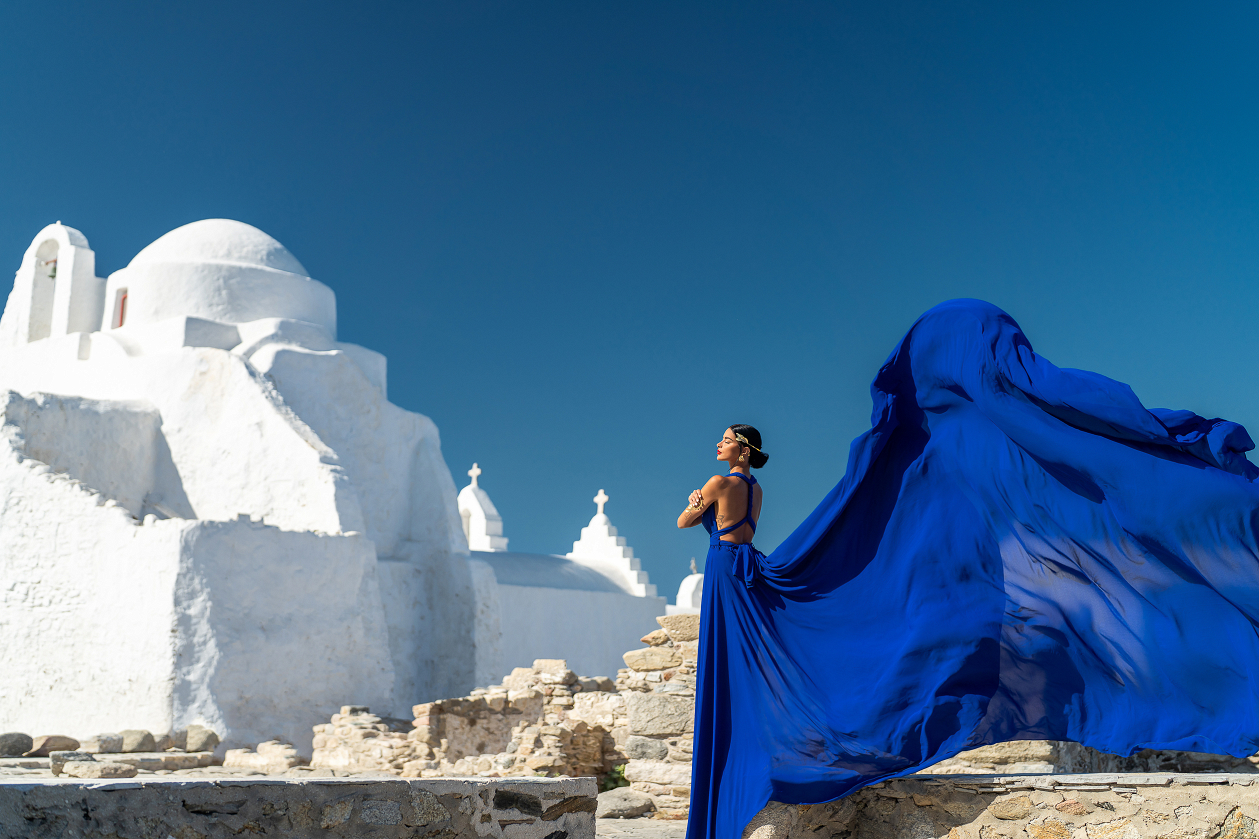 Embrace the enchanting beauty of Mykonos with a flying dress photoshoot at the Church of Panagia Paraportiani, where the elegant blue gown flows dramatically against the iconic white architecture and clear blue sky.